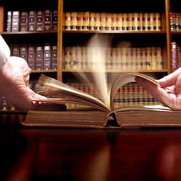 Despacho de procurador en Puertollano y Ciudad Real Dos manos intercambiando un libro sobre una mesa con estanterías de libros al fondo.